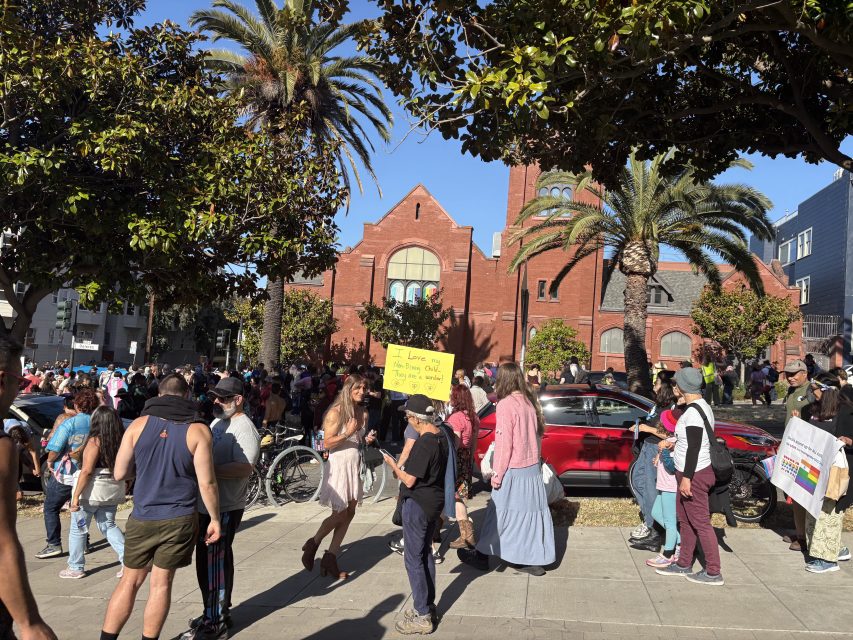 A crowd gathers outside a red-brick building with palm trees during a Pride event. One person holds a yellow sign; others walk or stand, some carrying bags or wearing costumes.