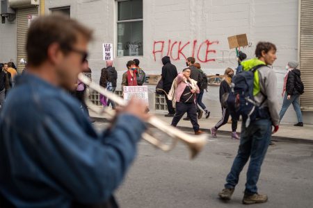 People walk past a building with protest signs and "FUCK ICE" graffiti; a trumpet player is blurred in the foreground, and one person holds a cardboard sign.