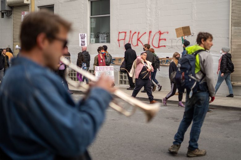 People walk past a building with protest signs and "FUCK ICE" graffiti; a trumpet player is blurred in the foreground, and one person holds a cardboard sign.