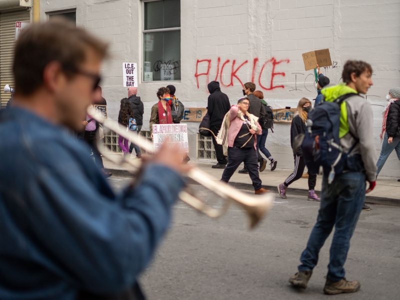 People walk past a building with protest signs and "FUCK ICE" graffiti; a trumpet player is blurred in the foreground, and one person holds a cardboard sign.