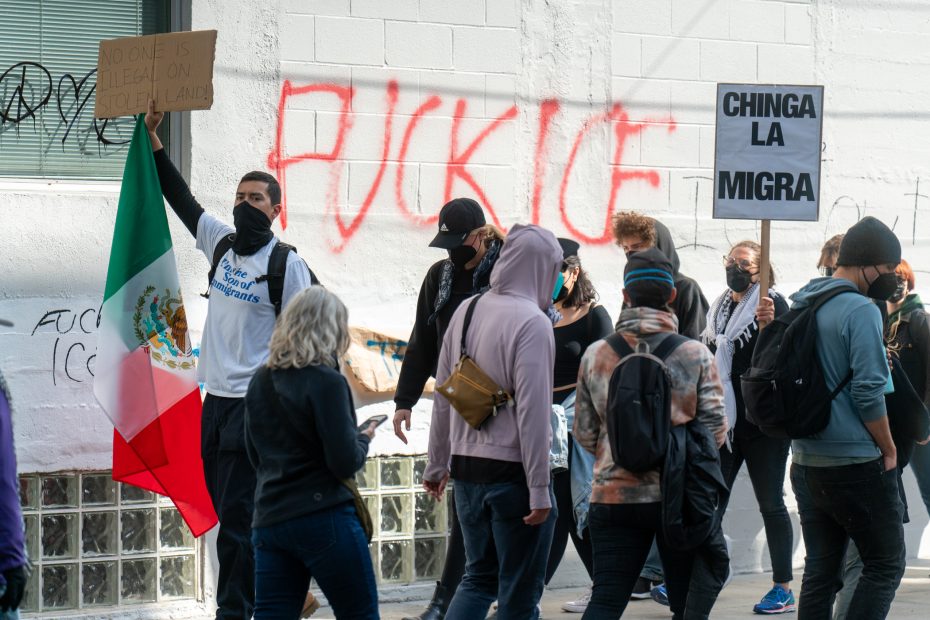 A group of people protest outdoors; one holds a Mexican flag, others hold signs, and red graffiti is visible on a white wall in the background.