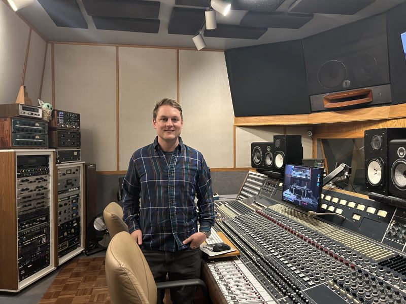 A person stands in a recording studio next to a large mixing console, surrounded by audio equipment and monitors.