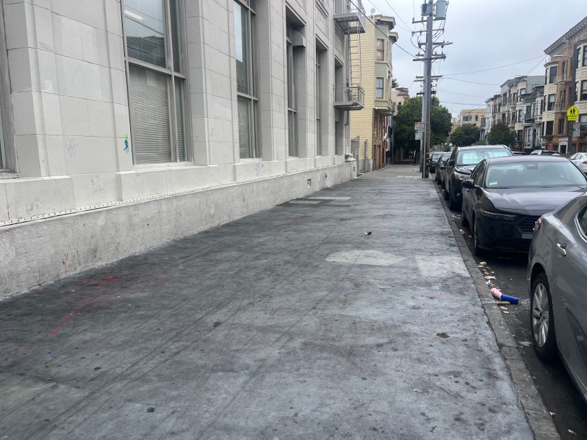 A city sidewalk runs alongside a gray building. Parked cars line the street, and scattered litter is visible on the pavement.