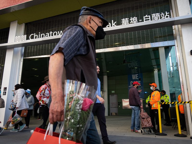 An older man carrying flowers and a gift bag walks past the entrance of Chinatown metro station, while others enter or exit and staff in orange vests stand nearby.