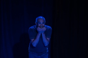 A man stands under blue stage lighting with his hands pressed to his mouth, expressing a nervous or anxious emotion.