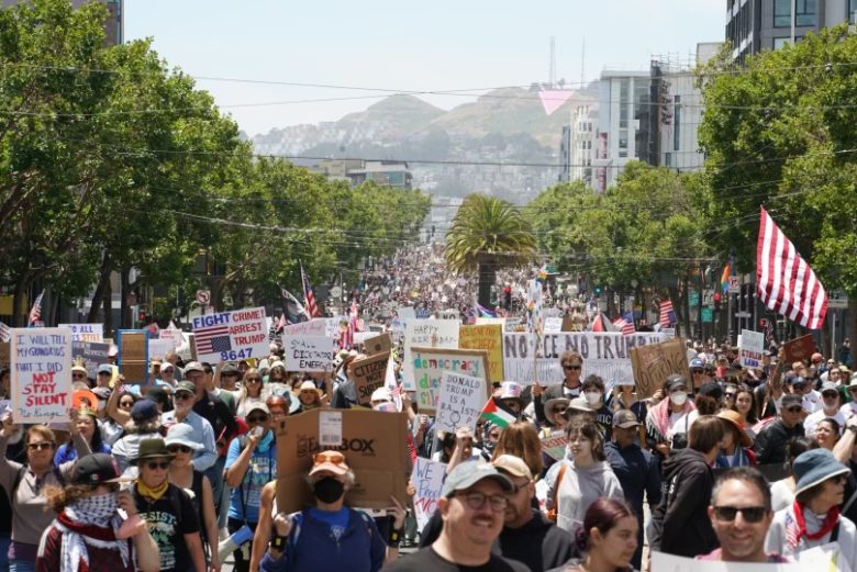 A large crowd of people marches down a city street holding protest signs, with trees lining the protest route and hills visible in the distance.