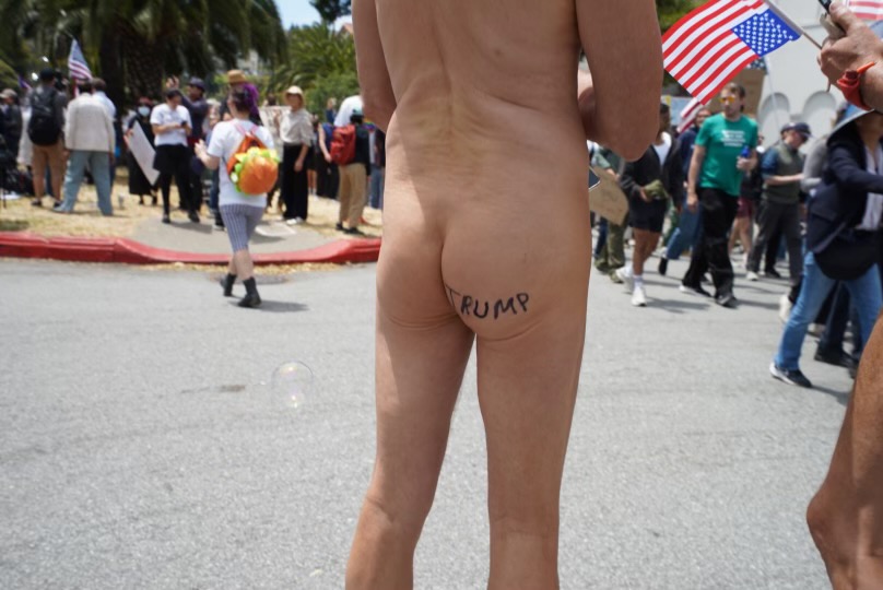 A nude person stands with "TRUMP" written on their buttocks at a public gathering; a crowd with flags is visible in the background.