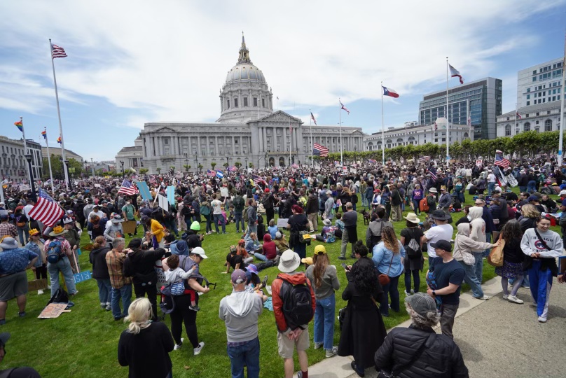 A large protest crowds the lawn in front of a government building, with flags, signs, and banners visible under a partly cloudy sky.