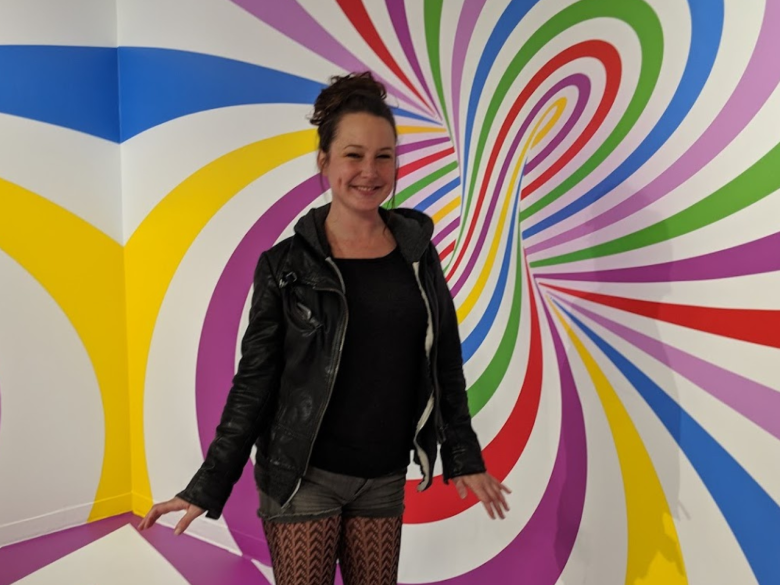 Johanna Luddy stands smiling in front of a colorful, swirling, optical illusion mural with patterns in blue, yellow, pink, green, red, and purple.
