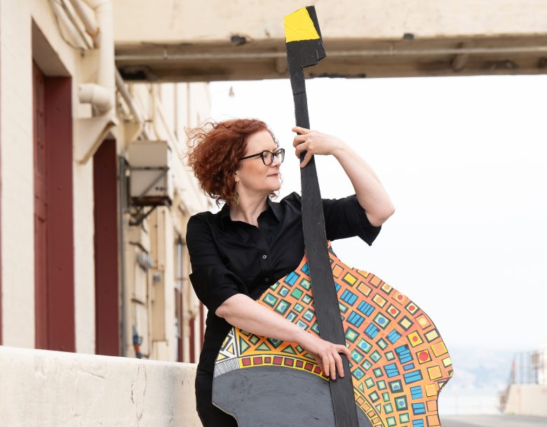 A woman with red curly hair and glasses holds a large, colorful, abstract guitar cutout while standing outdoors near a concrete structure.