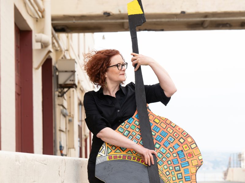 A woman with red curly hair and glasses holds a large, colorful, abstract guitar cutout while standing outdoors near a concrete structure.