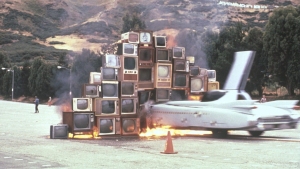A car crashes into a pyramid-shaped stack of old televisions, some of which are on fire, with mountains and trees in the background.