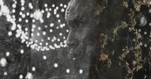 Black and white image of a person's face partially merged with a textured, rocky surface and blurred circular lights in the background.