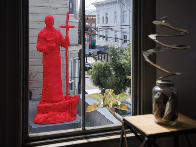 A large statue wrapped in bright red material stands outside near a building, viewed through a window with indoor decor in the foreground.