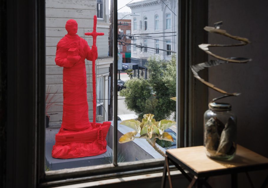 A large statue wrapped in bright red material stands outside near a building, viewed through a window with indoor decor in the foreground.
