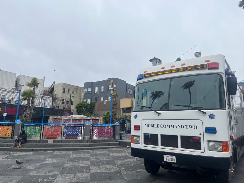 A police mobile command vehicle is parked in an urban square with colorful railings and buildings in the background on a cloudy day.