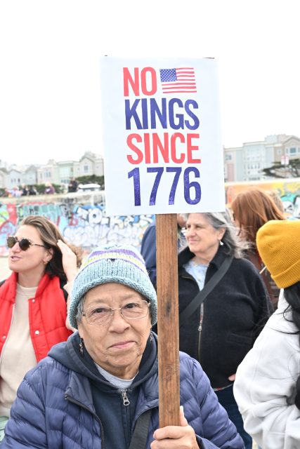 An older woman wearing glasses and a knit hat holds a protest sign that reads "No Kings Since 1776" with an American flag. Other people and buildings are visible in the background.