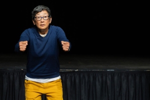 A person wearing glasses, a blue sweater, and yellow pants stands on stage with arms raised as if gripping an invisible steering wheel.