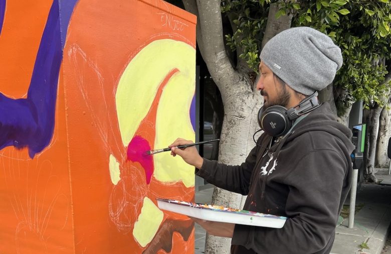 A man in a beanie and headphones paints a colorful mural on an orange utility box outdoors, using a brush and holding a palette.