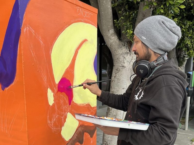 A man in a beanie and headphones paints a colorful mural on an orange utility box outdoors, using a brush and holding a palette.