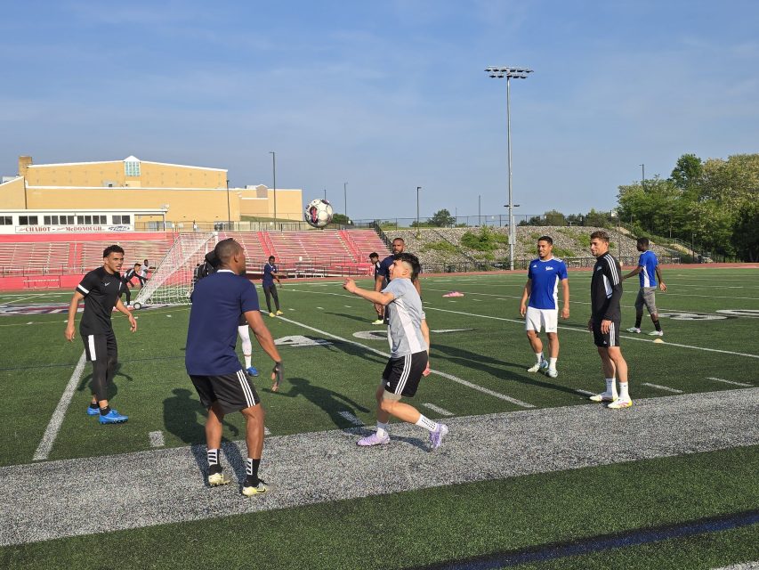 A group of men play soccer on an outdoor field, with one man preparing to head the ball while others watch or move nearby.