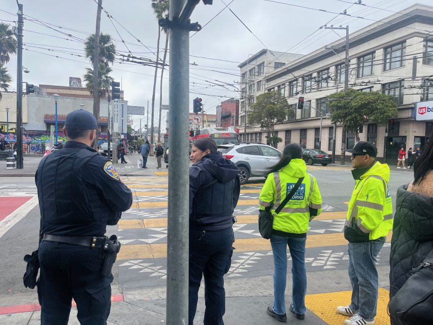 Two police officers and two people in yellow jackets stand at a city crosswalk, observing traffic and pedestrians on a cloudy day.