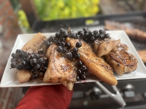 A white plate holds grilled fish fillets garnished with clusters of black grapes, with an outdoor grill visible in the background.
