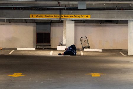 A blanket and a shopping cart sit in an empty, dimly lit parking garage with yellow signs indicating restricted height and no van parking.
