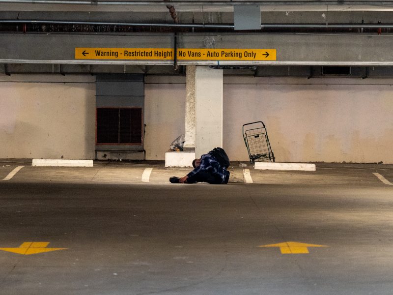 A blanket and a shopping cart sit in an empty, dimly lit parking garage with yellow signs indicating restricted height and no van parking.