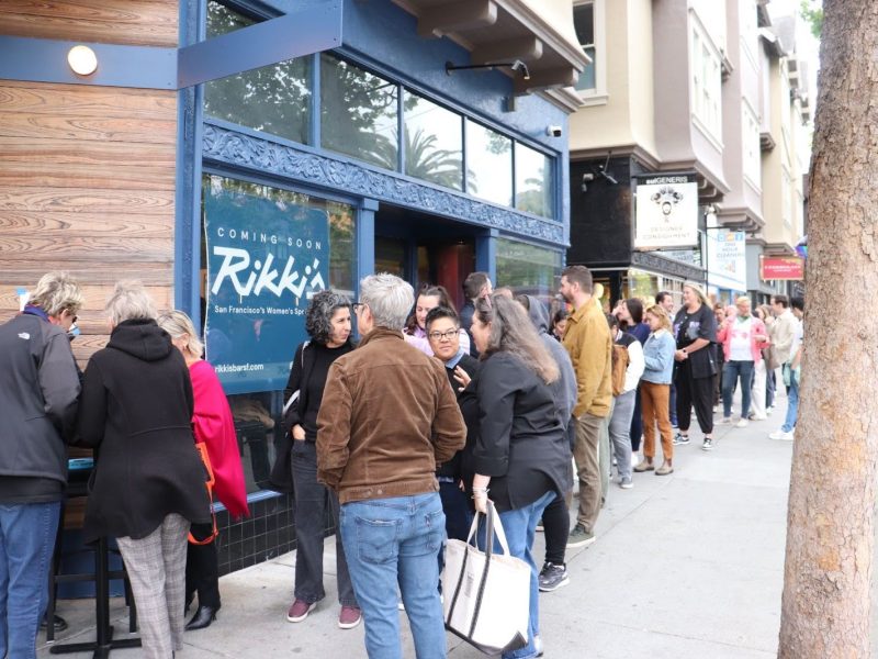 A long line of people stands on a sidewalk outside a storefront with a sign reading "Coming Soon Rikki's" during daytime.