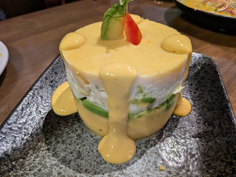 A layered potato and vegetable dish topped with sauce, garnished with a slice of avocado, a cherry tomato, and a basil leaf, served on a black rectangular plate.