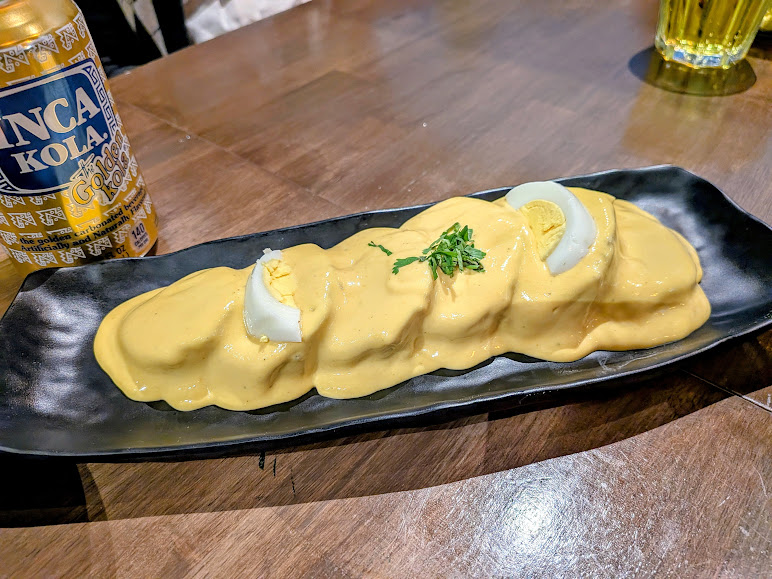 A black plate with four slices of potato topped with yellow sauce, garnished with chopped herbs and sliced boiled egg, next to a can of Inca Kola on a wooden table.