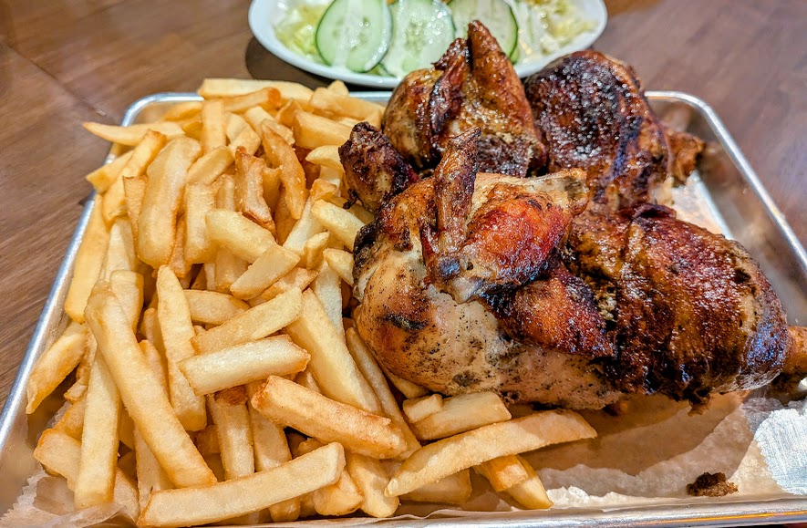 A tray with a serving of French fries and roasted chicken, with a plate of sliced cucumber and lettuce in the background.