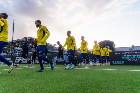 Soccer players in yellow and blue uniforms walk onto the field before a match, with the sun setting in the background.