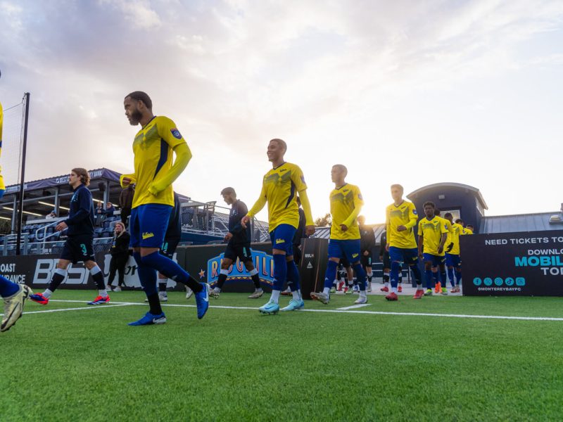 Soccer players in yellow and blue uniforms walk onto the field before a match, with the sun setting in the background.
