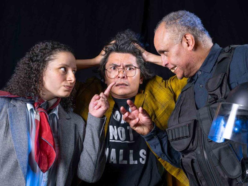Three people pose dramatically; one in a suit points accusingly, another in a yellow jacket looks stressed, and a third in riot gear gestures while holding a helmet.
