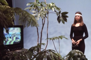 A woman in black workout attire and a white headband stands among plants beside a TV displaying colorful patterns against a blue background.