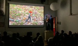 A group of people watch a projected nature image in a dark room while a person standing nearby plays a guitar.