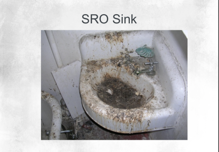A small, wall-mounted sink labeled "SRO Sink" is heavily stained and covered in dirt, grime, and debris, with a damaged green soap dish on the faucet.