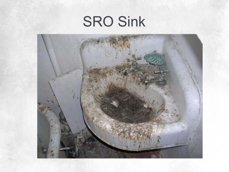 A small, wall-mounted sink labeled "SRO Sink" is heavily stained and covered in dirt, grime, and debris, with a damaged green soap dish on the faucet.