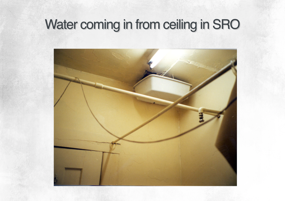 Ceiling in a room with visible water damage, a fluorescent light fixture, pipes, and an open panel showing water leaking from above.