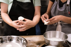 Two people wearing aprons shape dough with their hands over a wooden table, next to metal bowls containing liquid and ingredients.