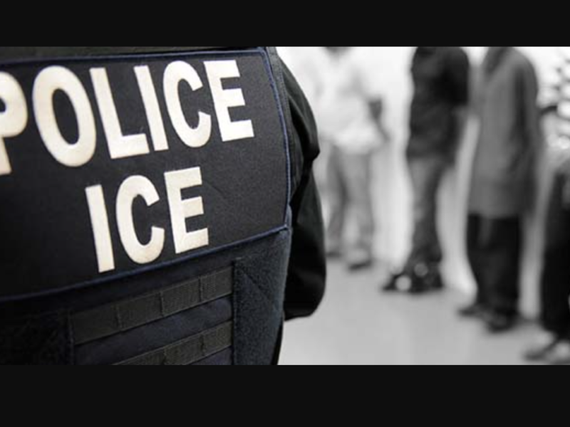 A person wearing a vest labeled "POLICE ICE" stands in front of a line of people against a wall.