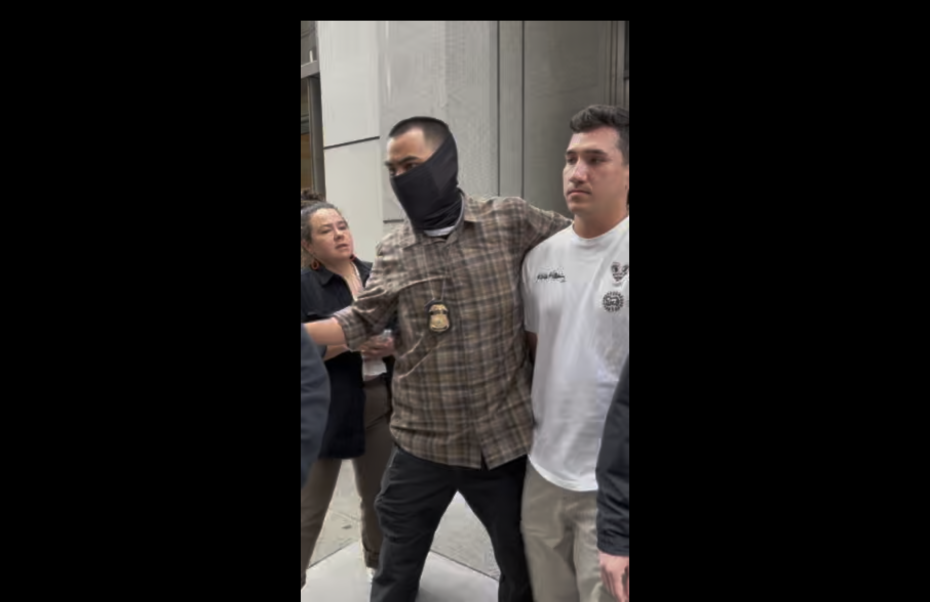 Two people escort a man in a white shirt on a city sidewalk; one escort wears a badge and mask, the other is behind them.