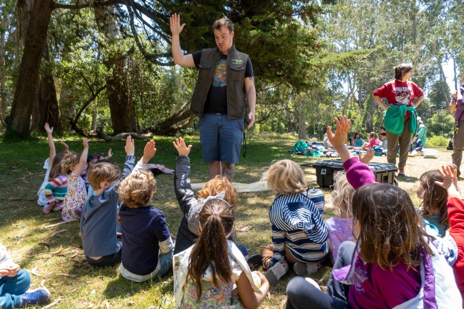 An adult stands and speaks to a group of children sitting on grass with hands raised in a wooded park area during the day.