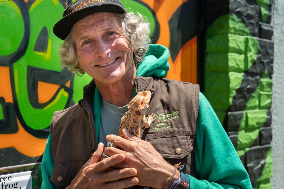 An older man with gray hair in a cap and vest smiles while holding a bearded dragon lizard, standing in front of a colorful graffiti wall.