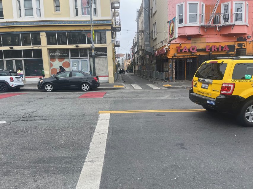 A city street intersection with parked cars, a yellow taxi, and a building with a "Taqueria" sign on the right side. The sky is overcast.