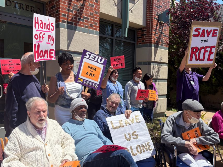 A group of people protest outside a building, holding signs with messages like “Hands off my Home,” “Save the RCFE,” and “Leave Us Alone.”.