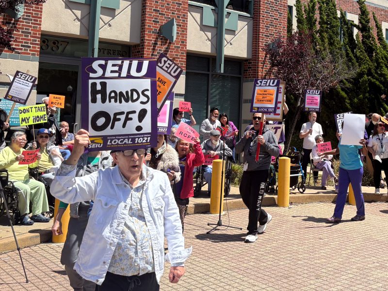 A group of people, some in wheelchairs, hold SEIU signs and protest outside a brick building. One sign reads “HANDS OFF!” in large letters.