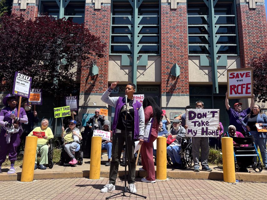 A person speaks at a microphone in front of a group holding signs that say "Don't take my home" and "Save the RCFE" outside a brick building.